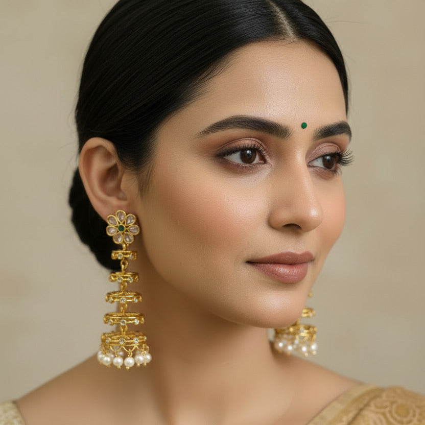 Gold earrings with floral designs and pearl accents held by a hand against a neutral background